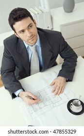 Architect Wearing Grey Suit Sitting At Office Desk, Writing Notes On Floor Plan, Looking Up. Overhead Shot.