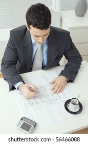 Architect Wearing Grey Suit Sitting At Office Desk, Writing Notes On Floor Plan. Overhead Shot.