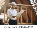 Architect wooden buildings. Man stands under frame wooden house. Man architect studies documentation or blueprints. Guy in protective helmet at construction site. Architect under timber framed house