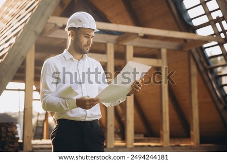 Architect wooden buildings. Man stands under frame wooden house. Man architect studies documentation or blueprints. Guy in protective helmet at construction site. Architect under timber framed house