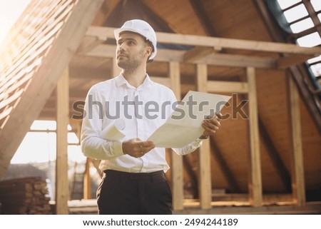 Architect wooden buildings. Man stands under frame wooden house. Man architect studies documentation or blueprints. Guy in protective helmet at construction site. Architect under timber framed house