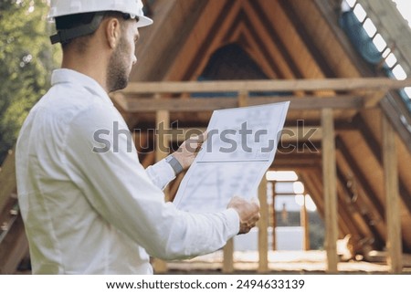 Architect wooden buildings. Man stands under frame wooden house. Man architect studies documentation or blueprints. Guy in protective helmet at construction site. Architect under timber framed house