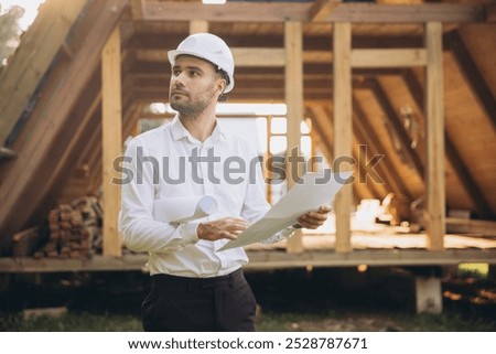 Architect wooden buildings. Man stands under frame wooden house. Man architect studies documentation or blueprints. Guy in protective helmet at construction site. Architect under timber framed house