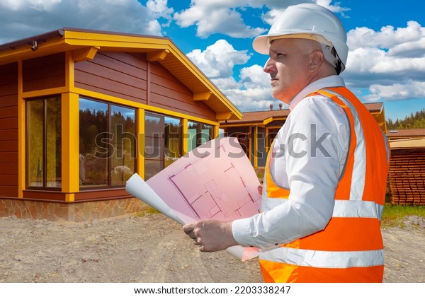 Architect\
working. Man architect examines blueprints of country cottage. Male\
employee of architectural bureau. Guy in orange vest in front of\
newly built house. Architecture company\
owner