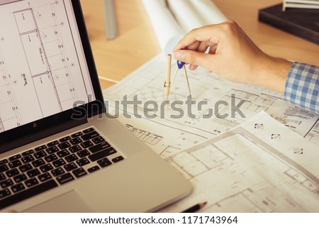 Architect working on blueprint in workplace with laptop and drawing compass.