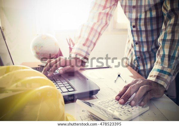 Architect working\
on calculator and laptop with construction plan on office desk\
under shinning light. film\
effect.