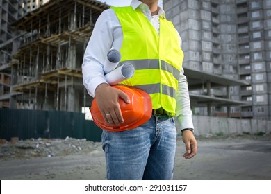 Architect in yellow safety jacket posing with red helmet at construction site