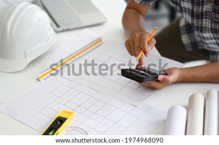 Architects are using a calculator to estimate the cost of house plans, house plans, safety helmet and office equipment placed on the desk.