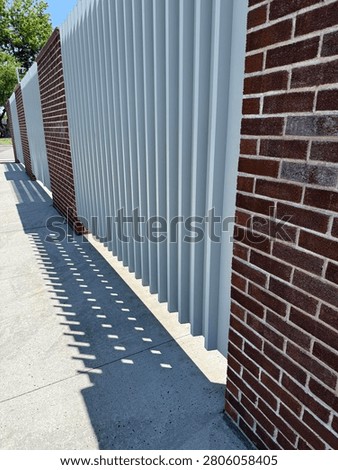 Architectural abstract of alternating red brick pillars and white vertical louvered panels with geometric shadows on a sunny sidewalk.