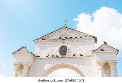 Architectural arch of temple gate with clock and cross at the top, surrounded by pigeons perched on the structure, under a clear blue sky with fluffy white clouds in the background