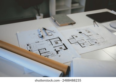 Architectural blueprints are spread out on a table with a pen, compass, and rolled up blueprints sitting on a table in an office setting - Powered by Shutterstock