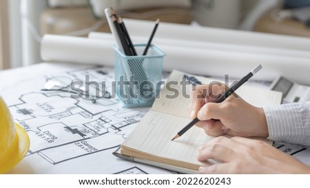 Architectural building design and construction plans with blueprints, young man is taking notes and planning a house or building with eraser, pen, pencil, tape measure, architect hat other equipment.
