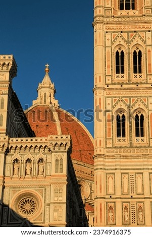 Architectural details of Florence Cathedral (Duomo di Firenze) showcasing its iconic red dome and ornate campanile bathed in golden sunlight against a clear sky.