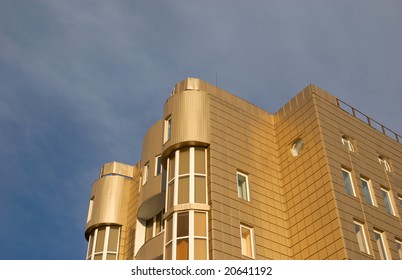 Architectural elegance: This contemporary structure ascends toward heavens, displaying metallic gold panels alongside expansive window features that create striking visual impact. A corner perspective emphasizes sculptural quality with rounded balconies and sleek lines, establishing sophisticated aesthetic. Natural illumination enhances warm golden tones, contrasting beautifully against cool blue sky adorned with cloud formations. Upward angle frames design elements to suggest growth and innovation, while highlighting intricate details and providing dimensional depth. Composition evokes feelings of awe and admiration through innovative urban design, perfect for real estate campaigns or architectural portfolios. Crisp clarity combined with nuanced interplay of light and materials makes imagery ideal for editorial content focused on metropolitan living.