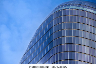 An architectural, low-angle photograph capturing the upper floors and distinct curved profile of a contemporary skyscraper. The building features a slick, grid-like facade composed of dark blue glazed glass window panels and polished steel framing lines. The curved building exterior sweeps upward against a bright, clear blue sky with soft wispy clouds. This clean, minimalist composition highlights innovative commercial engineering, futuristic design, and modern corporate environments.