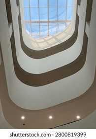 An architectural low-angle shot looking up through a multi-tier modern building atrium. The design features elegant, smooth, white curved walls and balconies illuminated by natural light coming from a large gridded glass skylight roof revealing a clear blue sky. Perfect for concepts related to interior design, modern architecture, minimalism, or urban development.