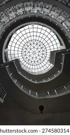 An architectural low-angle shot utilizing a symmetrical glass dome to create a compelling radial composition, framed by the rhythmic, curvilinear silhouettes of a multi-tiered spiral staircase.
