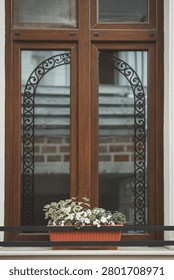 ​Premium architectural medium shot showcasing a classic European-style dark wood window frame in a beautiful vertical composition. The image captures two elegant glass panes decorated with traditional black wrought iron scrollwork stencils that form a perfect decorative arch. On the marble window sill, a charming terracotta plastic flower box is filled with blooming white impatiens (Busy Lizzie) and variegated green-and-white foliage, creating a soft, welcoming focal point. The glass subtly reflects a rustic brick and stone wall across the street, adding depth and an authentic urban story to the scene. This harmonious, warm composition is an exceptional commercial asset engineered for property real estate branding, southern European travel editorials, cozy home decor blogs, and lifestyle hospitality marketing.