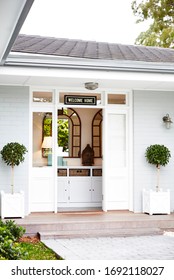 Architectural Photography Of A Grey Hamptons Style Home Facade With A Topiary Trees At The Entrance And A Bench Set And A Garden Bed, Front Door Open Showing A White Hall Stand And Mirror