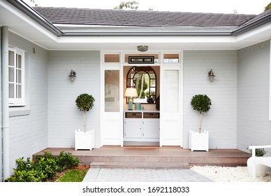 Architectural Photography Of A Grey Hamptons Style Home Facade With A Topiary Trees At The Entrance And A Bench Set And A Garden Bed, Front Door Open Showing A White Hall Stand And Mirror