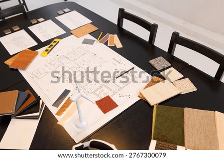 Architectural plan sitting on table in studio with veneer, pencils, spirit level and turbine model. Layout, materials, samples, tiles, woodgrain, palette, workspace