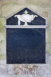 Architectural Slate Frame. 13th Century Carved Stonework, With Decorative Urn With Fabric Detail, Resting On A Stone Plinth, With A Dark Slate Area And Empty Space For Text.,