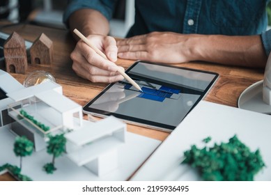 Architecture designer holds a pencil pointing to the 3D home office drawing on the tablet while thinking and considering detail in the building, Interior design and landscape around.