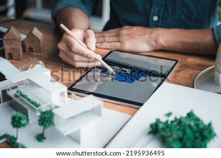 Architecture designer holds a pencil pointing to the 3D home office drawing on the tablet while thinking and considering detail in the building, Interior design and landscape around.
