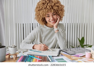 Architecture and interior design concept. Happy curly haired female architect draws sketches for future apartment chooses colors for building decoration smiles broadly poses at home at desktop. - Powered by Shutterstock