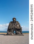 The Arctic Henge in Raufarhöfn, Iceland, features stacked stone arches and a conical top, set against a clear blue sky and ocean backdrop.