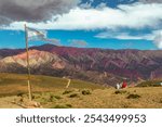 The Argentine flag flies as two people contemplate the imposing panoramic view of the multicolored Hornocal mountains, Quebrada de Humahuaca, Argentina,