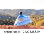 Argentinian man seated atop a mountain, proudly holding the Argentine flag, surrounded by a mountainous landscape. Celebrating Argentina