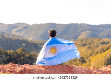 Argentinian man seated atop a mountain, proudly holding the Argentine flag, surrounded by a mountainous landscape. Celebrating Argentina's Independence Day. - Powered by Shutterstock