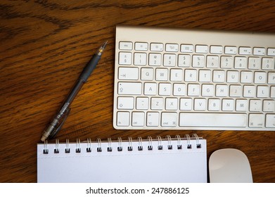 An Arial View Of An  IMac Wireless Keyboard, Mouse, Pen, And Desk Pad On An Oak Wood Desk