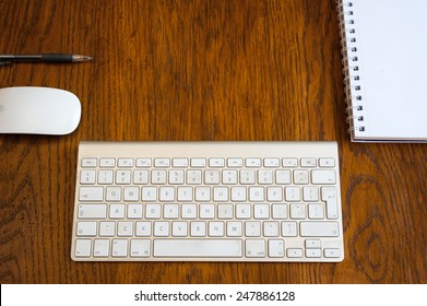 An Arial View Of An  IMac Wireless Keyboard, Mouse, Pen, And Desk Pad On An Oak Wood Desk