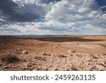 Arid Arizona landscape with rocky foreground, Meteor Crater in center, and distant mountains under a sky of white clouds and blue patches. Remote natural beauty captured in late afternoon light.