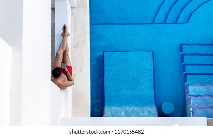 Ariel Shot Of An Attractive And Athletic Young Man Lounging Next To A Swim Up Pool In A Hotel Texting Or Using A Cellphone Or Mobile Phone On A Luxury Holiday Or Vacation In Mexico