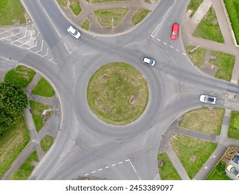 Ariel shot taken from drone showing a roundabout with light traffic. Showing road markings, grass, paths and cars.