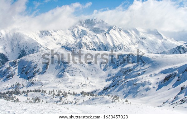 Ariel view of snowy mountains and blue sky in Bansko\
resort, Bulgaria. Majestic high mountain peaks covered with snow\
and airy white clouds. Nature wallpaper with winter holiday and\
skiing resort. 