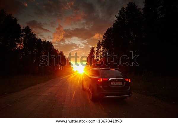 Arkhangelsk region, Russia - August 2017: Mitsubishi\
Outlander car rides on dirt road to horizon, against the bright\
sunset sky with the rays of the sun and the black silhouette of\
trees. Freedom 