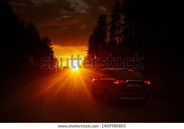Arkhangelsk region, Russia - August 2017: Mitsubishi\
Outlander car rides on dirt road to horizon, against the bright\
sunset sky with the rays of the sun and the black silhouette of\
trees. Freedom 