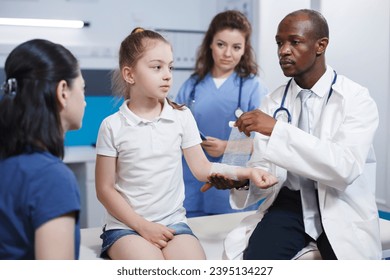 Arm of girl patient being meticulously wrapped with a sterile bandage by multicultural medical personnel. Caucasian youngster accompanied by her mother is being treated by doctor and nurse. - Shutterstock ID 2395134227