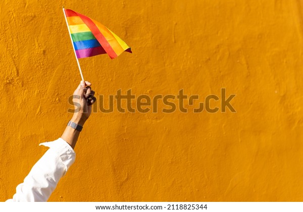 arm of an unrecognizable black Latin\
person waving holding a gay LGBT flag on a yellow background. LGBT\
rainbow ribbon pride tape symbol. Stop\
homophobia.