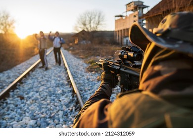 An Armed Soldier On The Railway In Uniform With A Modern Rifle Aimed At Two People. A Soldier Guarding The Border Stops Illegal Border Crossing