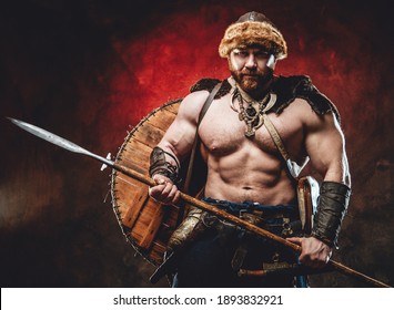 Armed with spear muscular and shirtless scandinavian barbarian with shield on his back poses in dark red background looking at camera.