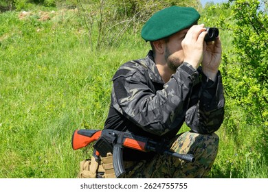 Armed special forces commando soldier in green beret scouting through binoculars hidden behind foliage during reconnaissance in active combat warzone