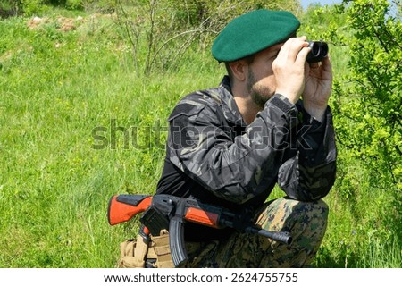 Armed special forces commando soldier in green beret scouting through binoculars hidden behind foliage during reconnaissance in active combat warzone