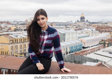 An Armenian girl with flowing long brown hair in a checkered shirt sits on a rooftop