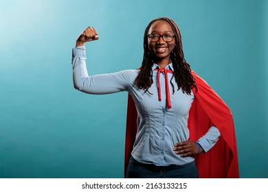 Arms Muscle Flexing Brave Superhero Woman Posing Strong And Tough For Camera On Blue Background. Justice Defender Wearing Mighty Hero Red Cape While Expressing Empowerement. Studio Shot.