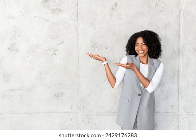 With arms pointing aside, a joyful African-American businesswoman presents an invisible object, her engaging smile and open stance suggesting a friendly invitation to potential opportunities - Powered by Shutterstock